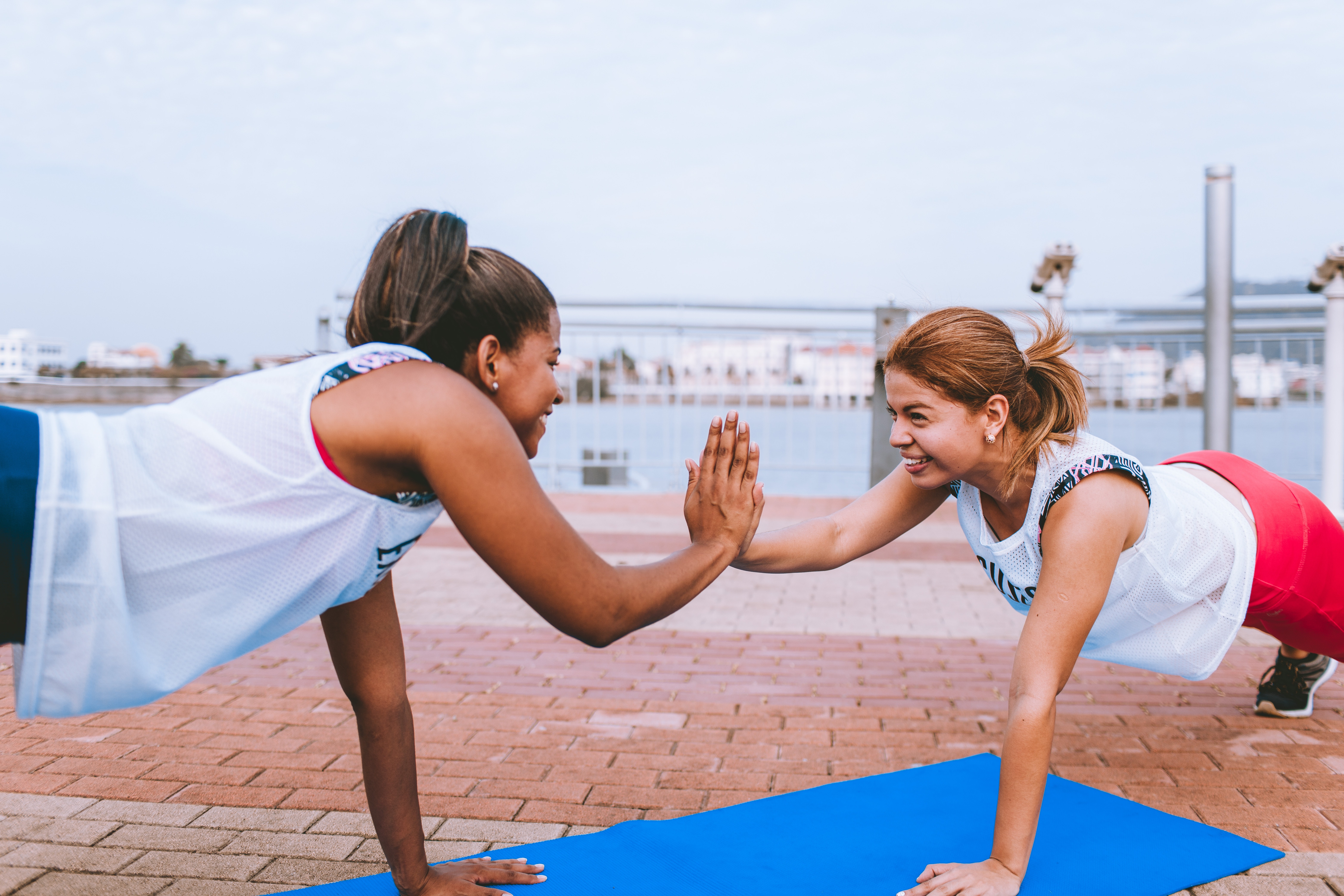 Two women are facing each other and smiling. They are performing a plank and high fiving on a pier.