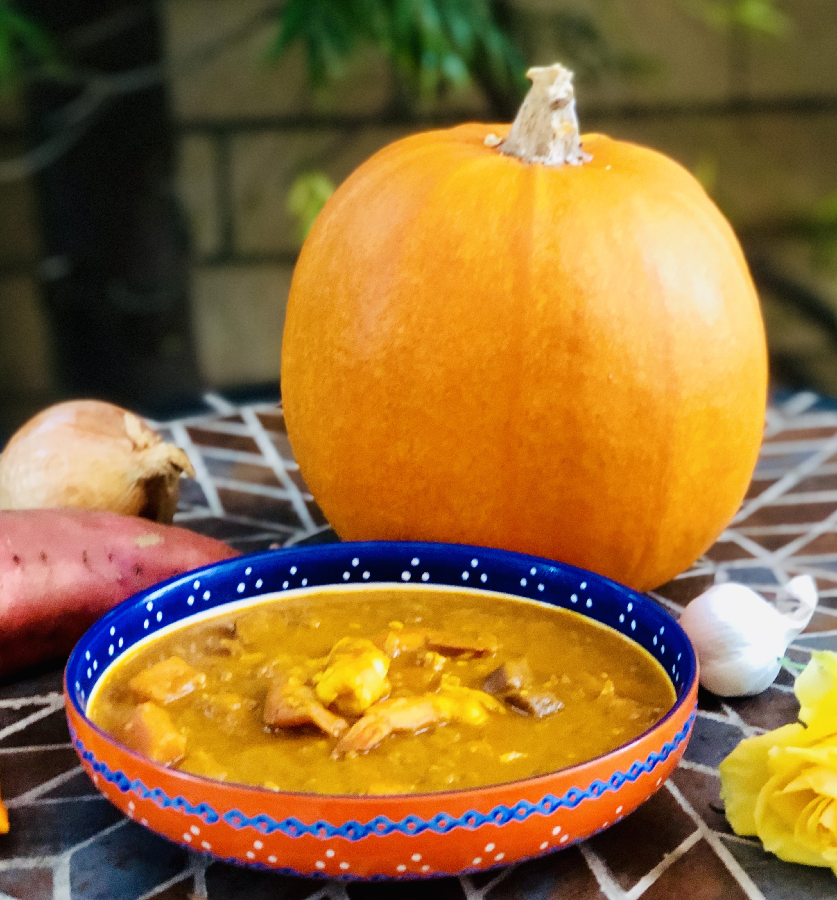 Bowl of orange curry on an outdoor table with a large orange pumpkin, purple sweet potato, onion, head of garlic, yellow and yellow rose