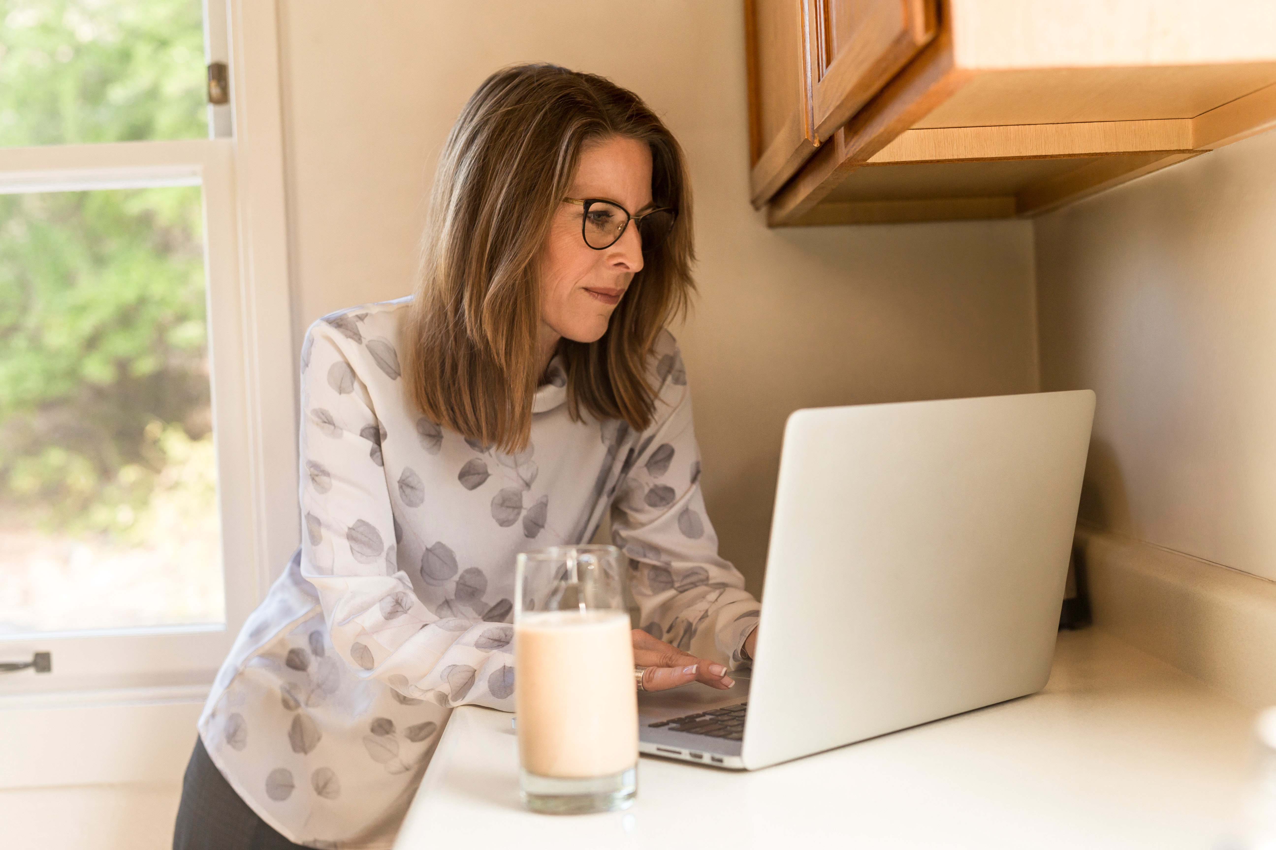 Woman standing typing on laptop with glass of milk on counter