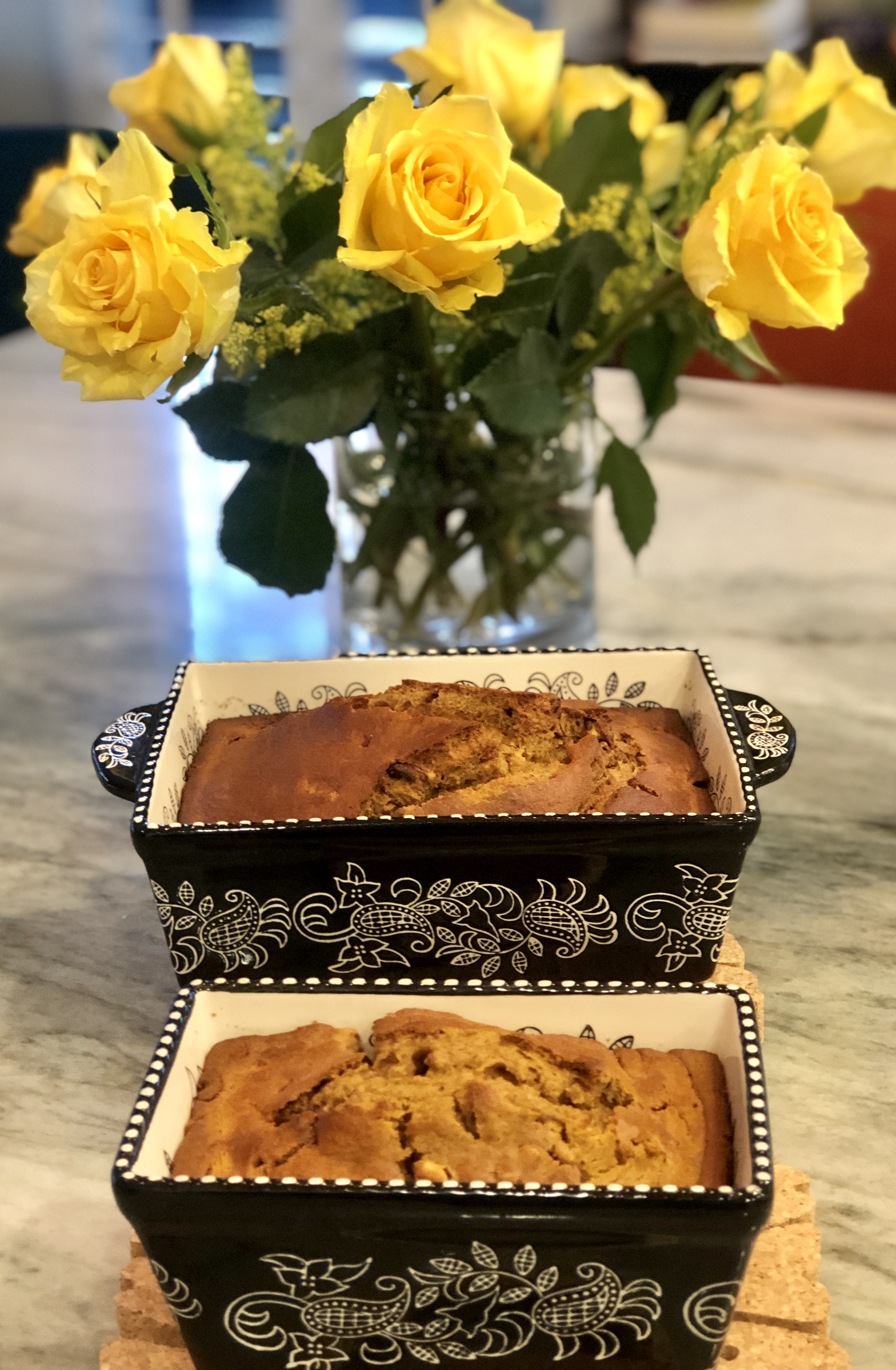 Vase with bright yellow roses and 2 loaves of golden brown pumpkin bread in Spanish style ceramic pans on Molé Mama's marble kitchen counter.