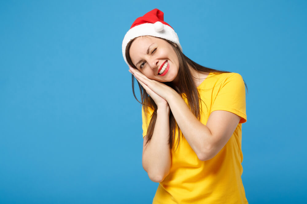 A woman in a bright yellow shirt and red Santa hat places her head against her hands, indicating sleepiness.; all against blue background