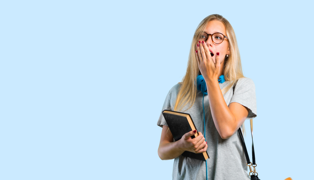 Student girl with glasses yawning and covering wide open mouth with hand. Sleepy expression on blue background