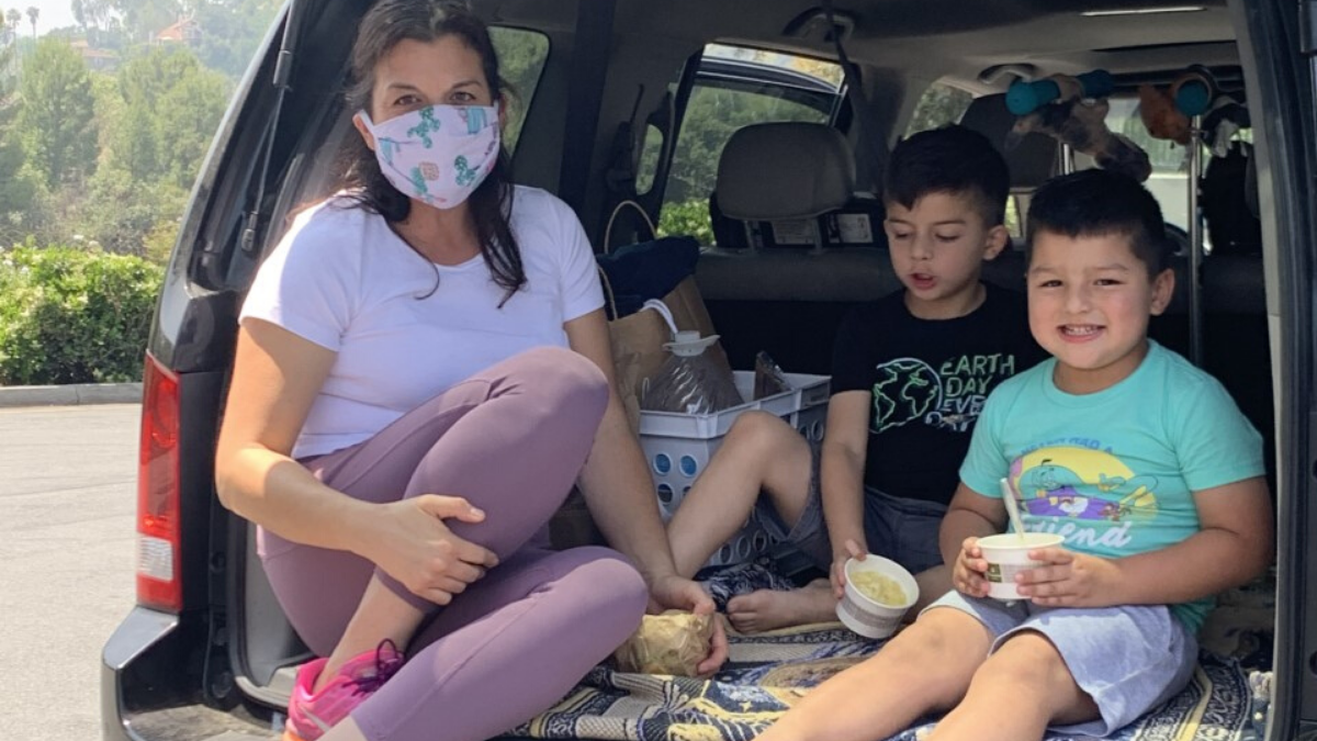 Photo of parent wearing a mask with two sons having lunch sitting on their trunk outside