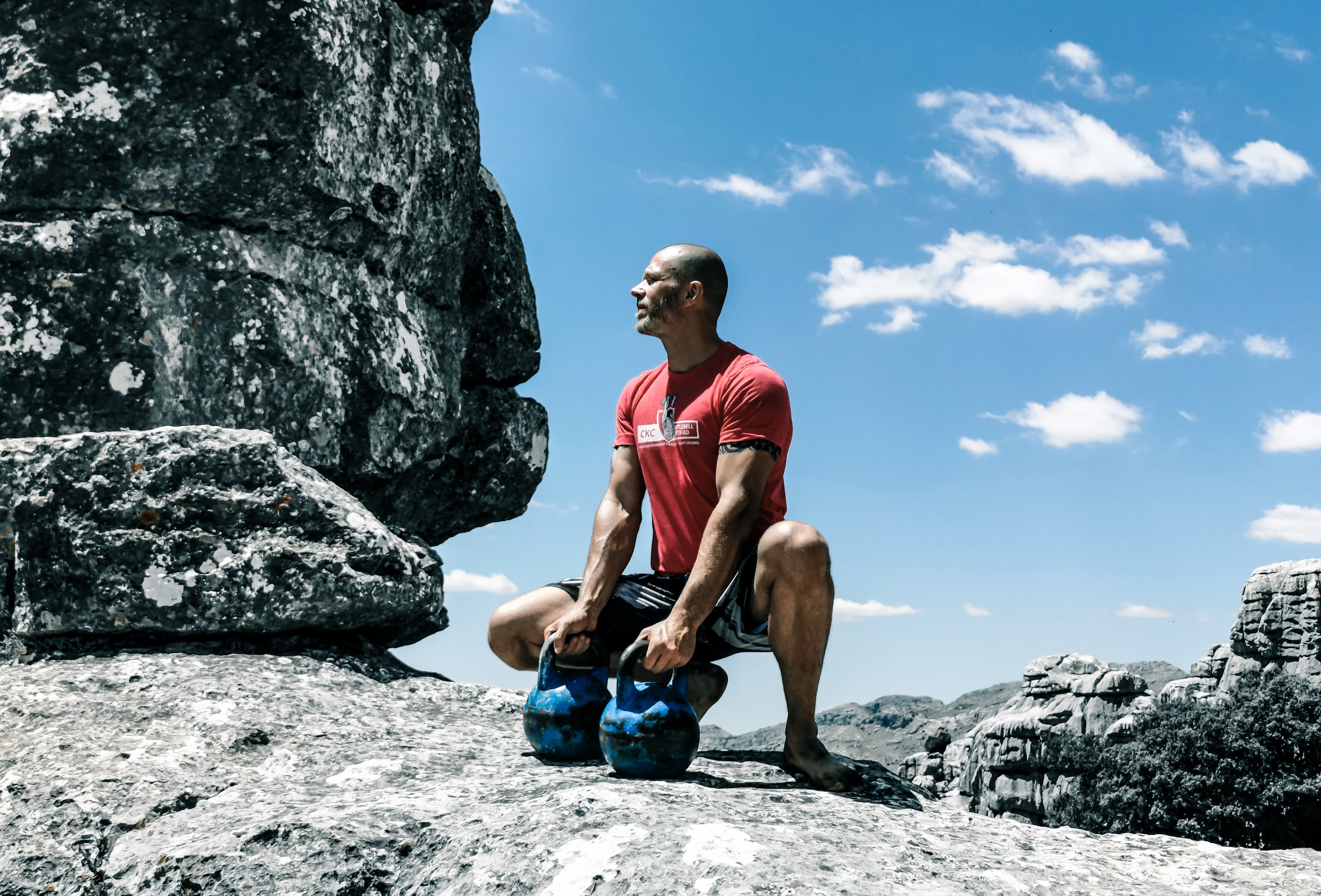Man in red shirt squatting down on the edge of a cliff holding 2 blue kettle bell weights