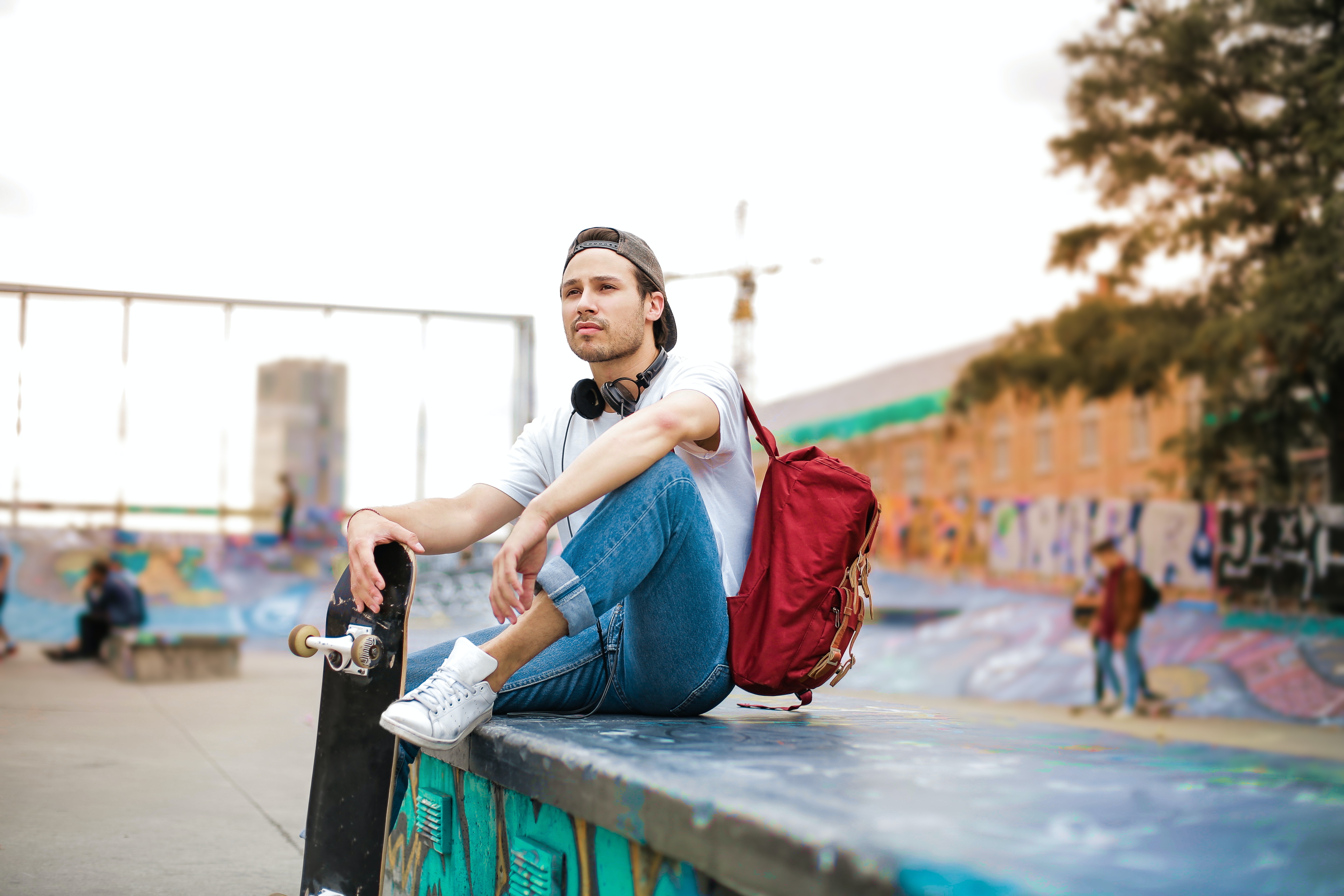 Man in shite shirt, blue jeans and red backpack, sitting down holding a skateboard