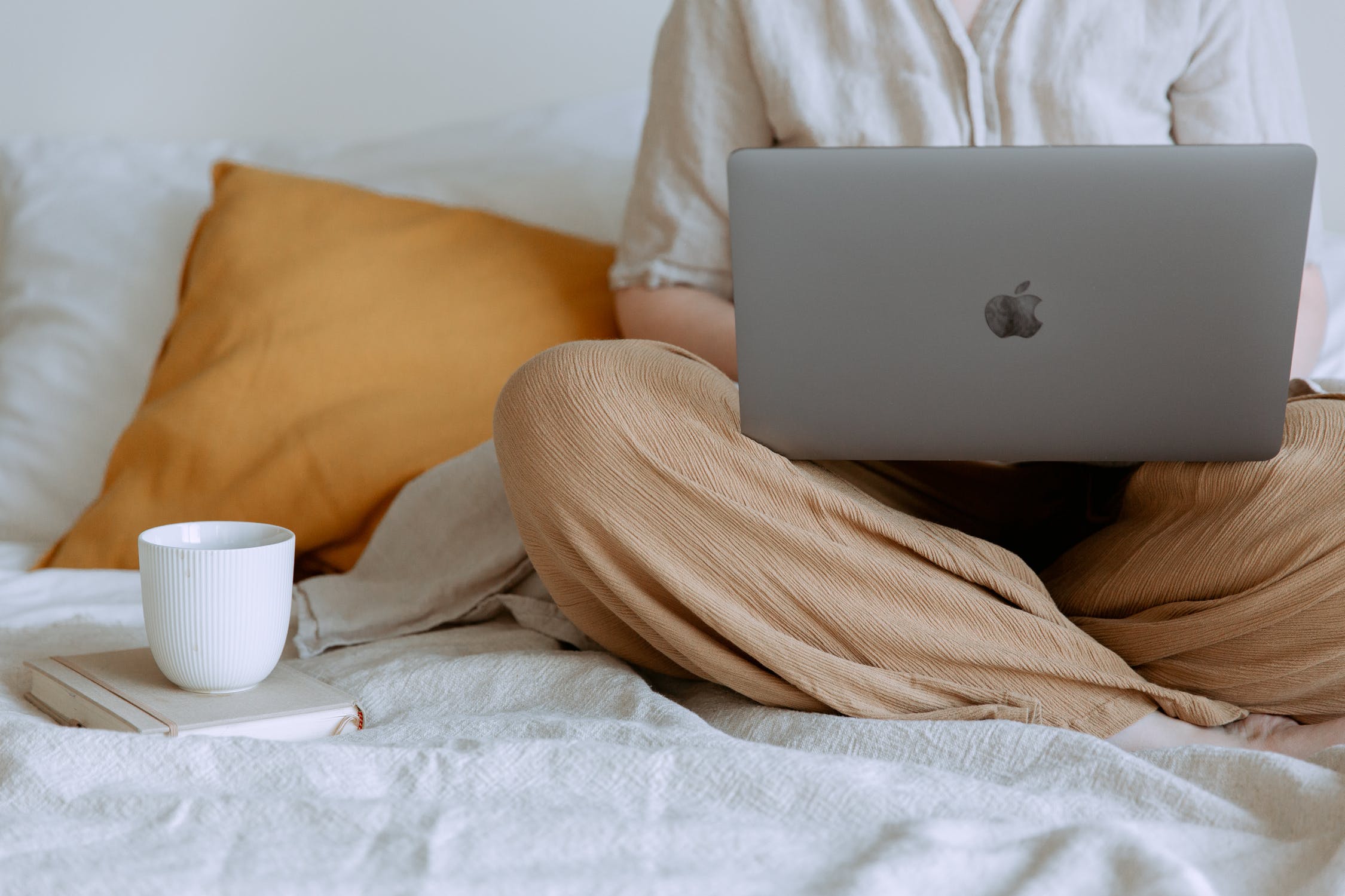 Woman sitting with her legs crossed on white linens with a laptop and coffee