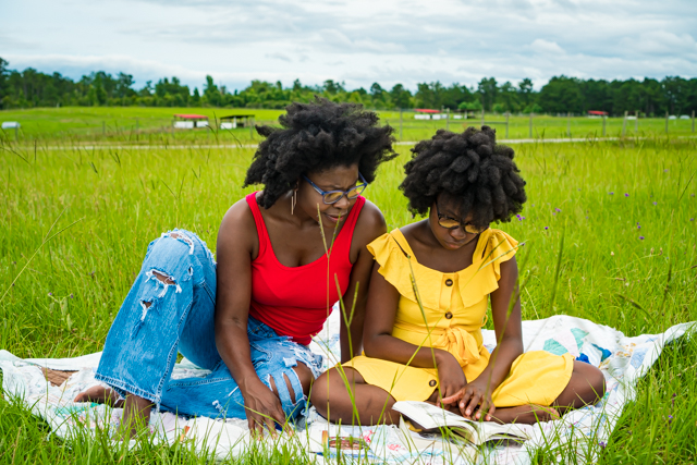 Girl sitting on a blanket in a grass field reading with her mother