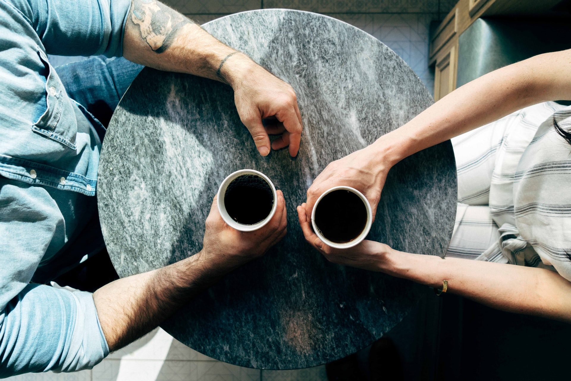 Two hands holding two cups of coffee across a table.
