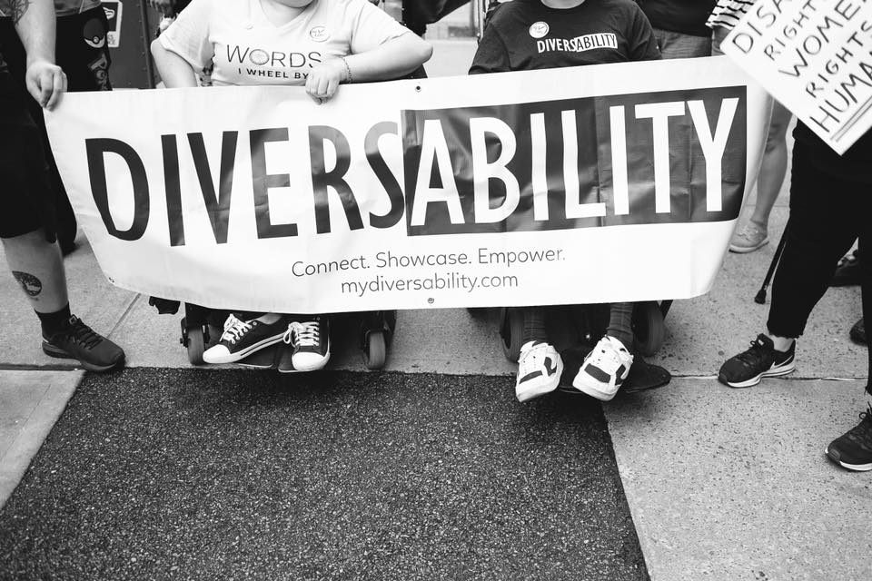 A black and white photograph of Diversability community members holding the Diversability sign. Two of the members are sitting in wheelchairs. It is a close up image of the banner, with the community member’s heads cropped out.