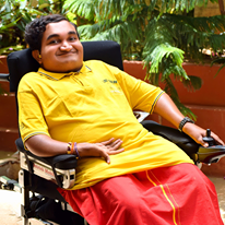 Dark skinned man with brown eyes and black hair. He is wearing a bright yellow shirt and red bottoms. He is sitting in a wheelchair. He is smiling.