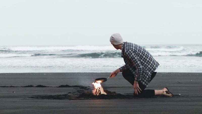 A man cooking on a camp fire in front of a beach