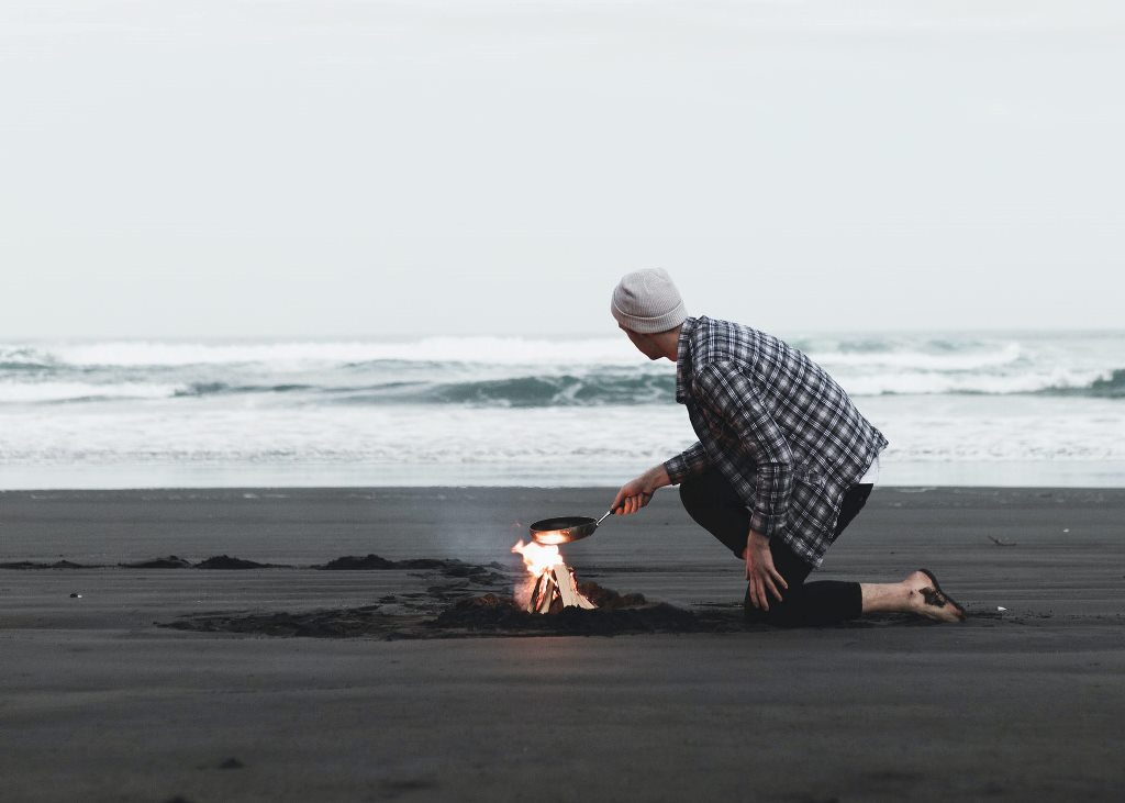 A man cooking on a camp fire in front of a beach