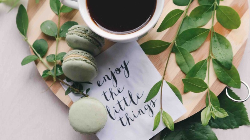 cup of coffee on a wood board, with green leaves, macarons and a napkin that says "Enjoy the little things."