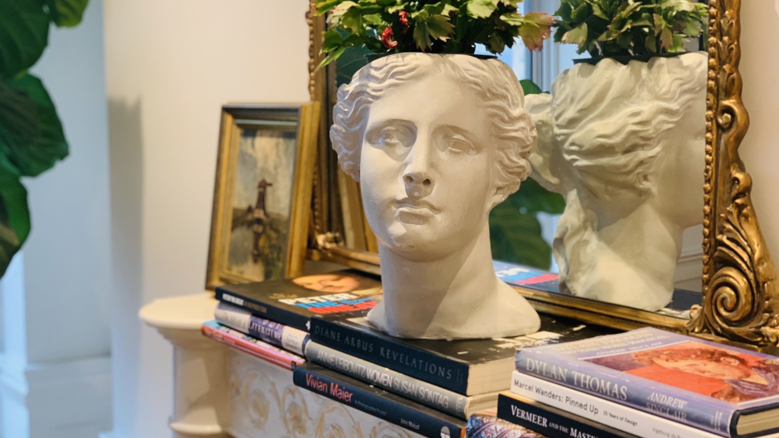 Planter bust on top of stack of coffee table books in front of mirror.