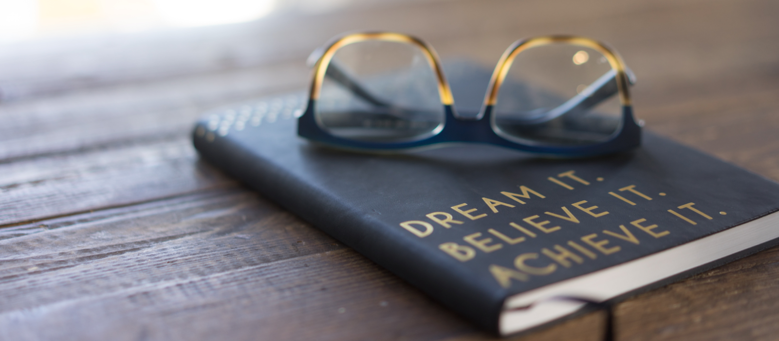 Glass on top of journal reading "Dream it. Believe it. Achieve it." Photo for Unsplash by Carolyn Christine.