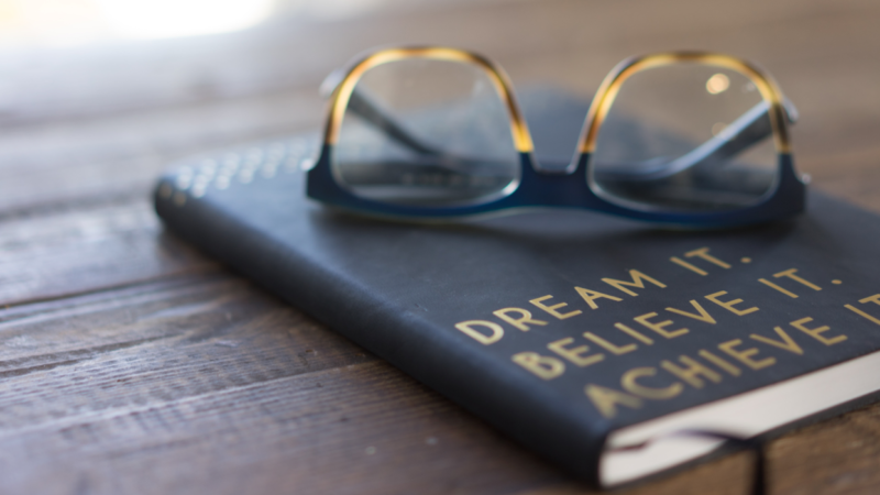 Glass on top of journal reading "Dream it. Believe it. Achieve it." Photo for Unsplash by Carolyn Christine.