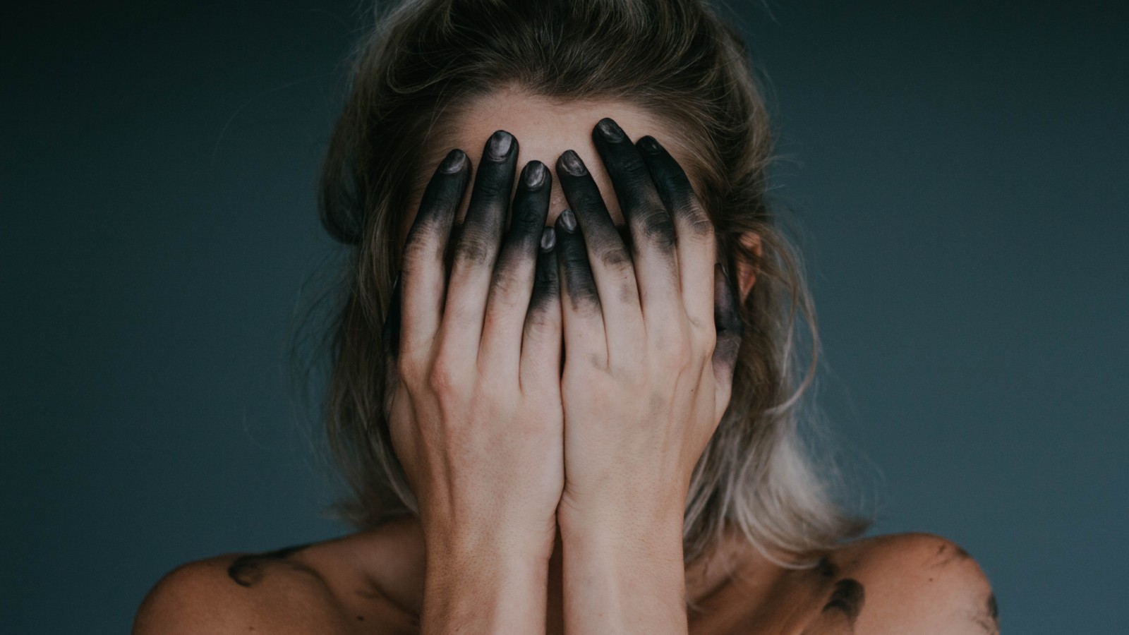 Woman covering her face. Tips of her fingers are covered in charcoal to represent feelings of burnout.