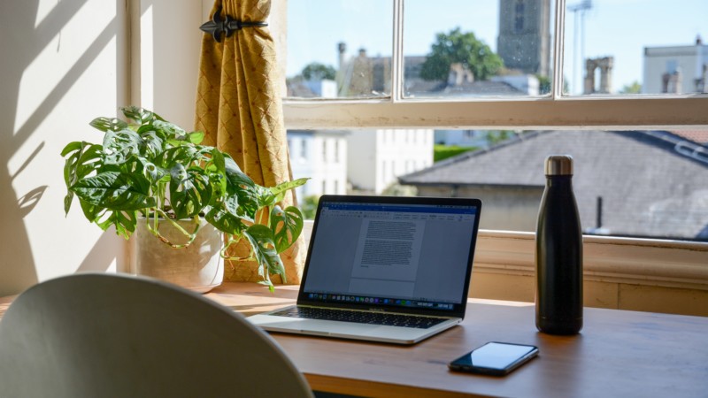 Computer sitting on desk next to a plant, the window is open and the sun is shining
