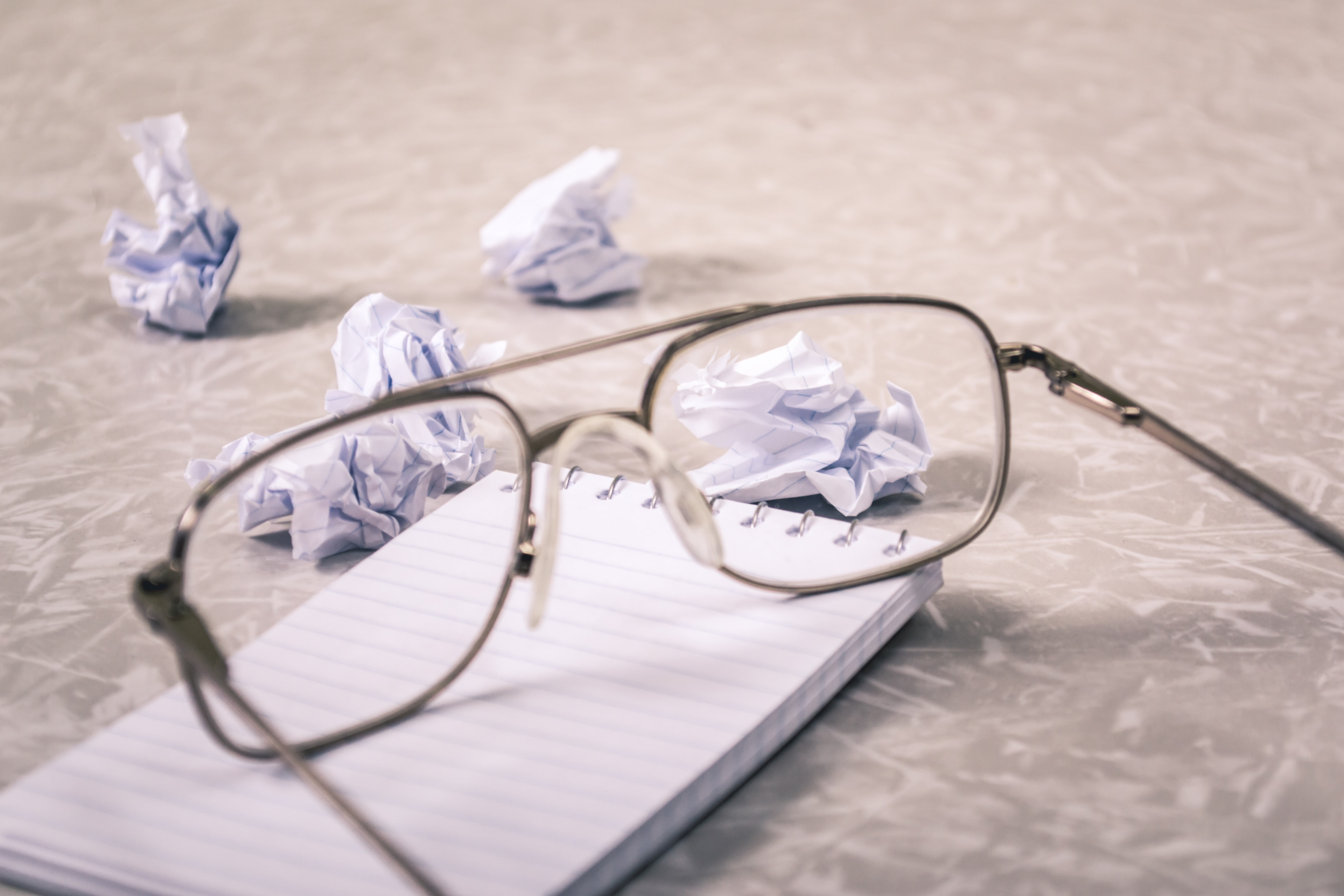A pair of glasses left on a notepad. Littered with balls of crumpled up paper. 