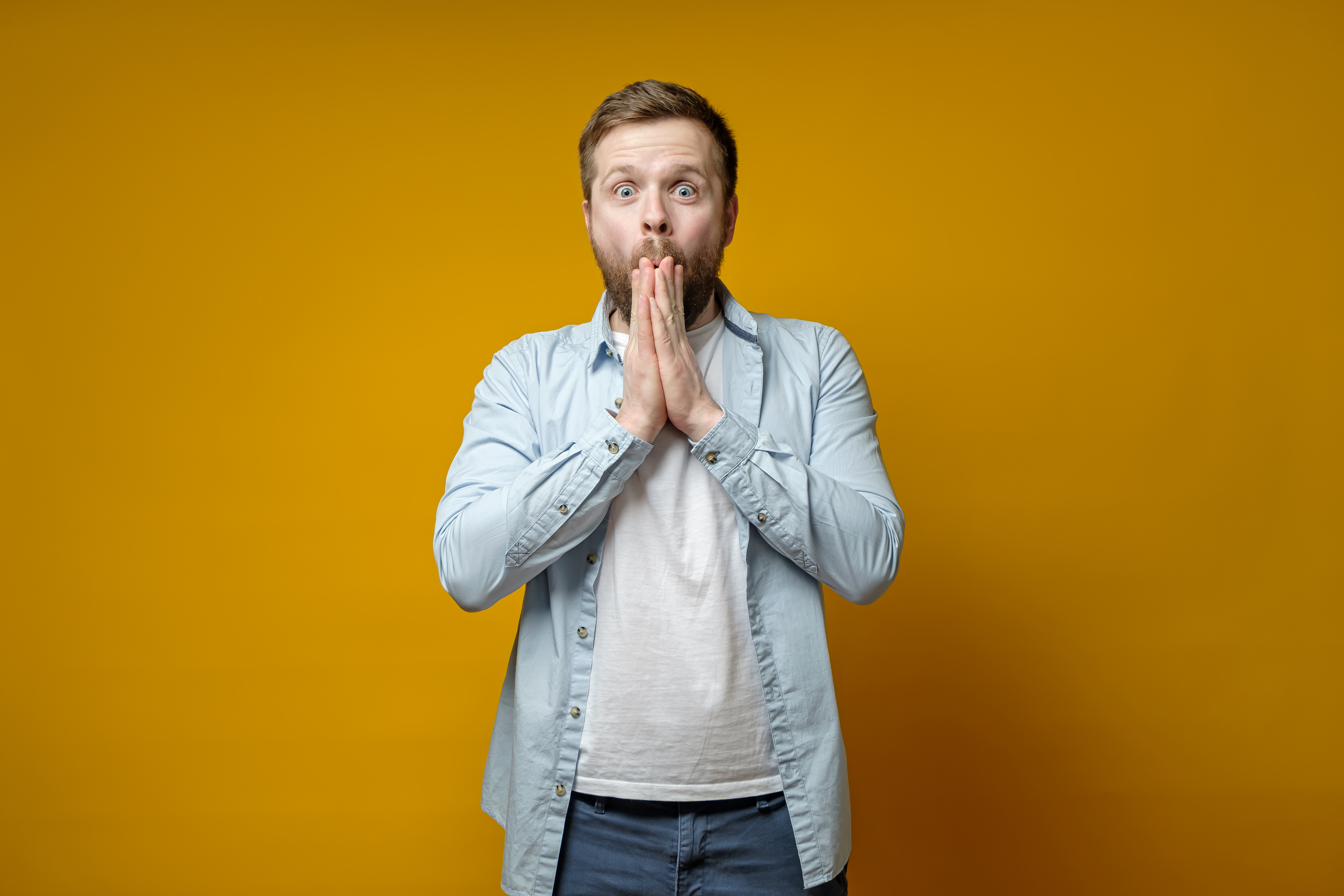 Caucasian, bearded man is pleasantly surprised by an unexpected surprise, he is happy and in a stupor, holds hands at mouth and looks with big eyes at the camera. Isolated on a yellow background.