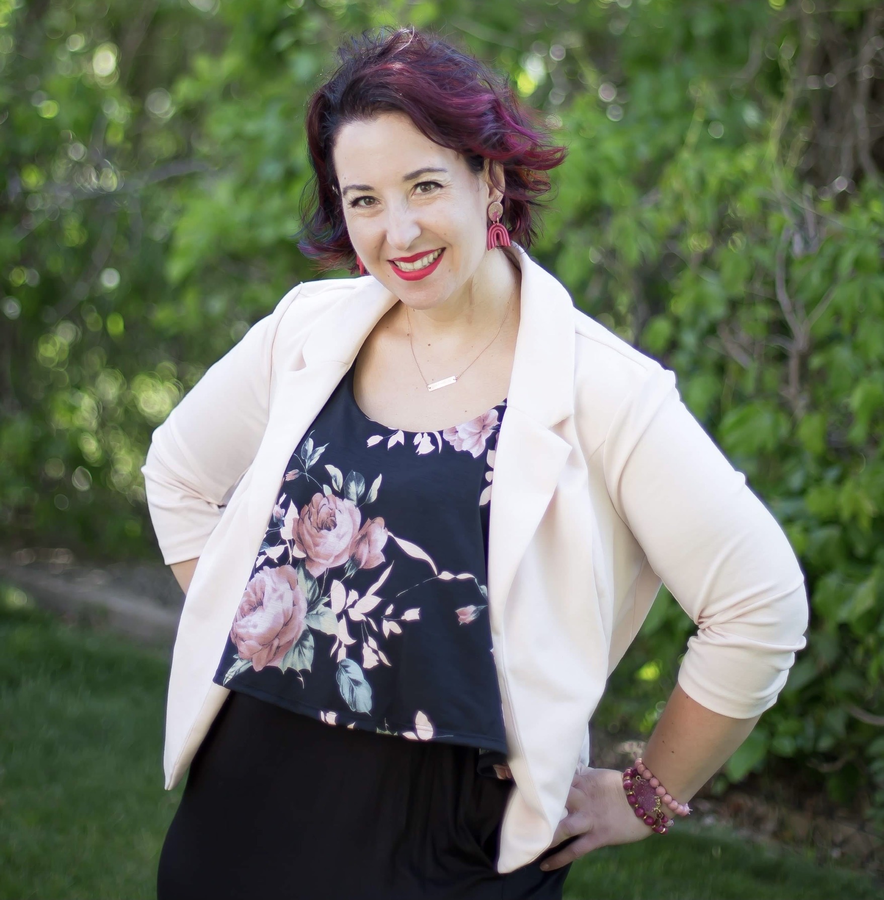 A white woman and outspoken advocate with multiple sclerosis wearing a black dress with a floral top and pink blazer while smiling in her back yard.