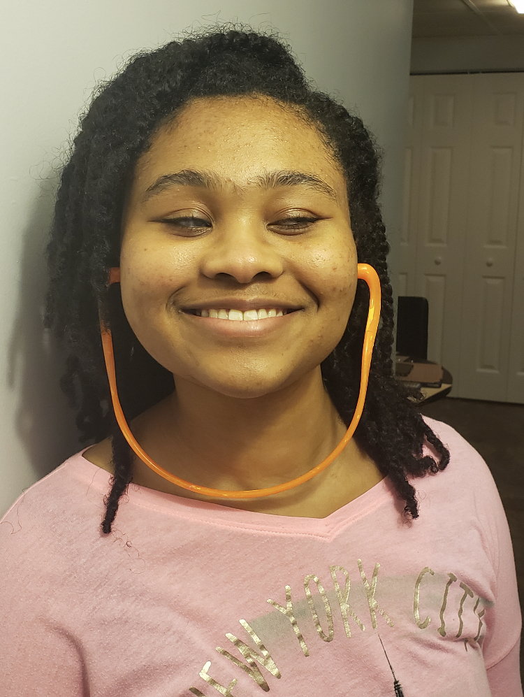 Jemma, an African American woman with natural hair smiles a toothy grin as she is standing against a wall wearing orange hearing bands and a pink shirt with gold foil that says ‘New York City’ on the font.
