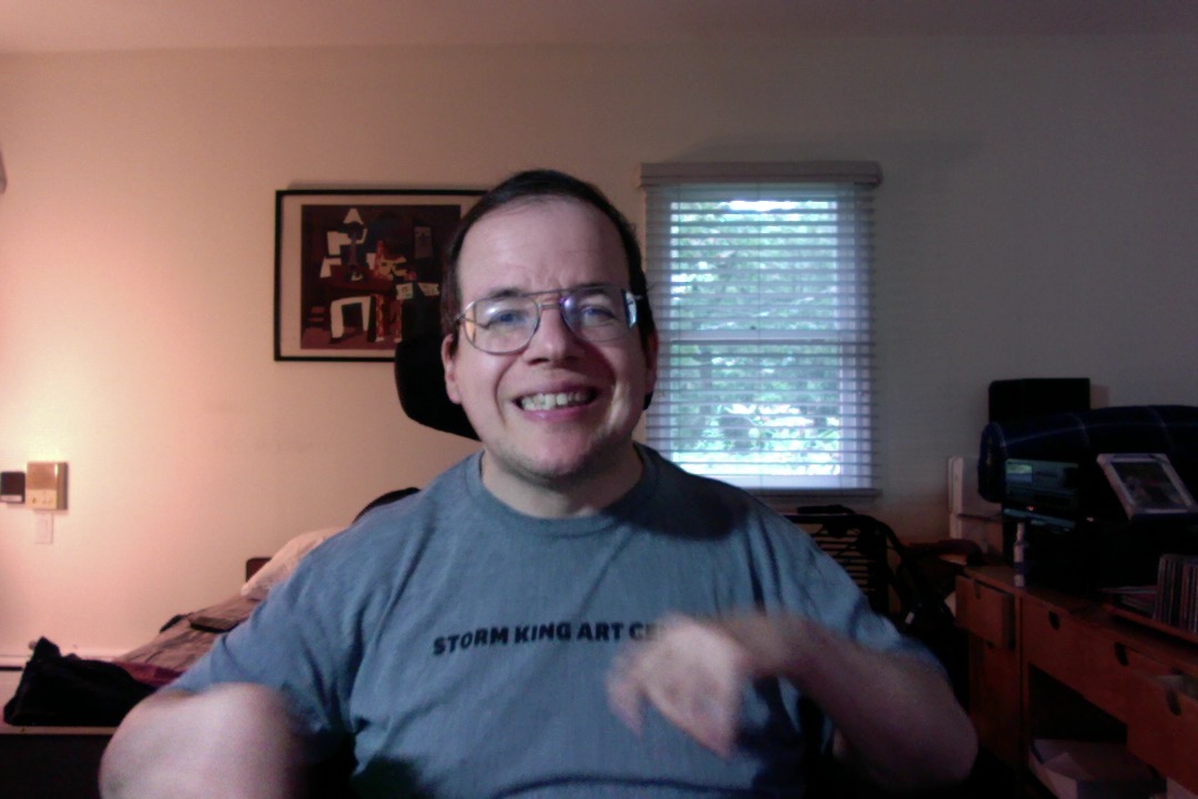James Michels, a smiling 50 year old man with brown eyes, glasses, thinning brown hair that has grey patches, and it is visually apparent that he has spastic triparesis and is right dominant with my left arm somewhat contracted. He is wearing a dark gray T-shirt with Black writing reading "Storm King Art Center".