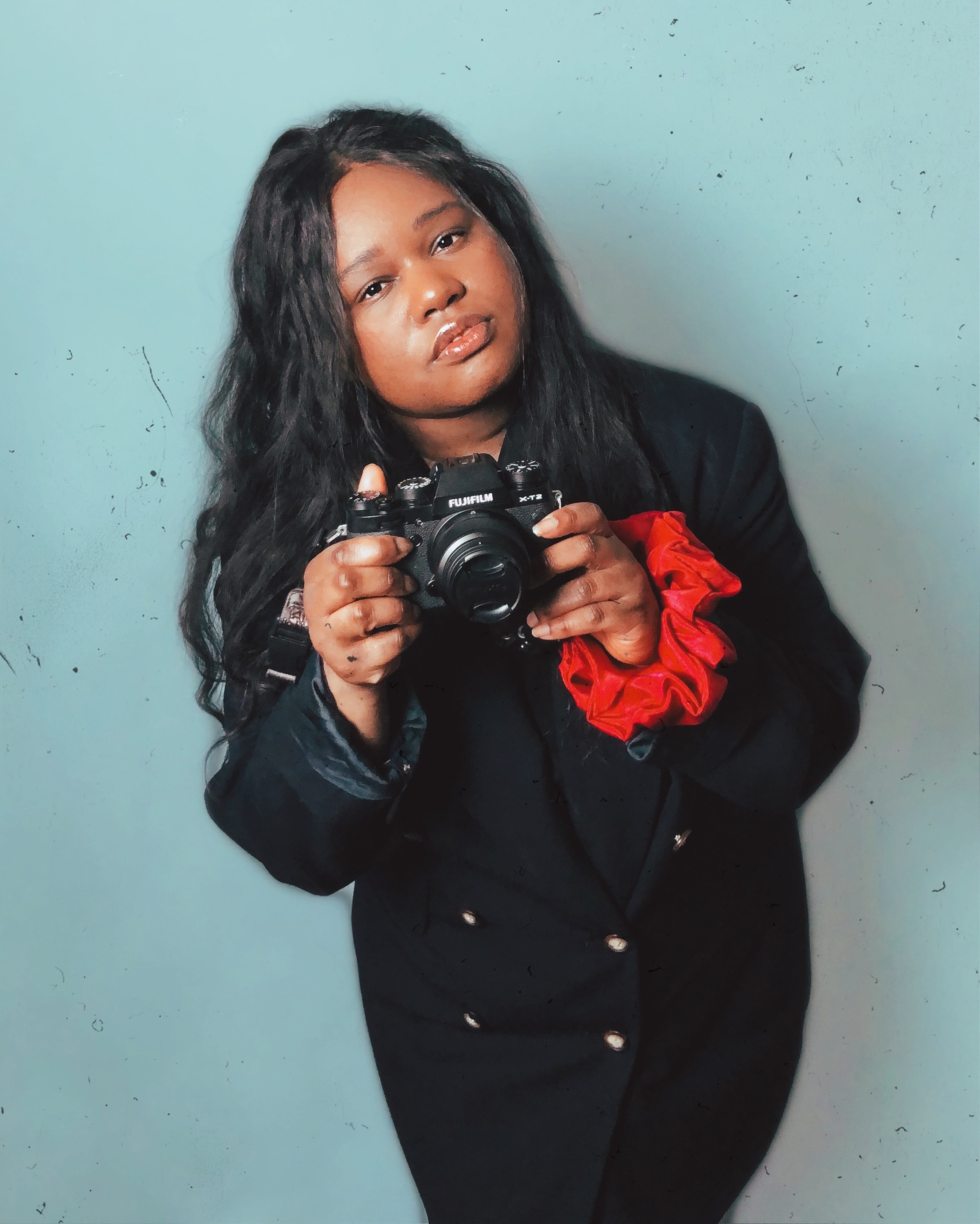 Jacquelyn is a black woman with long black hair. She is holding a digital camera and wearing a red scrunchie.
