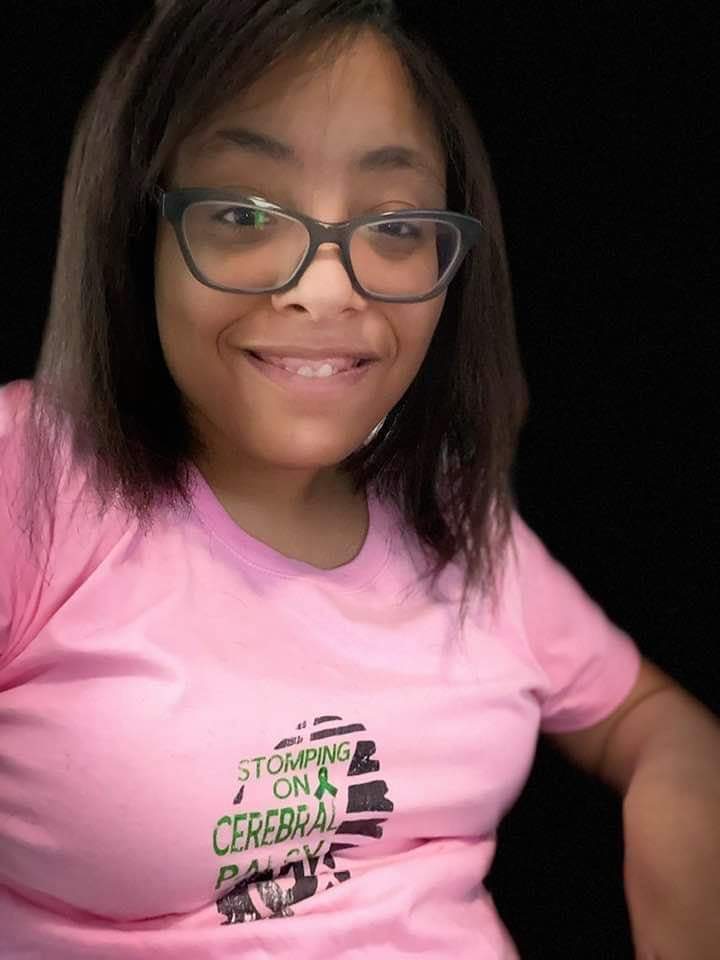 An African-American Latina with a black background wearing glasses and a pink t-shirt with the stomping on CP logo