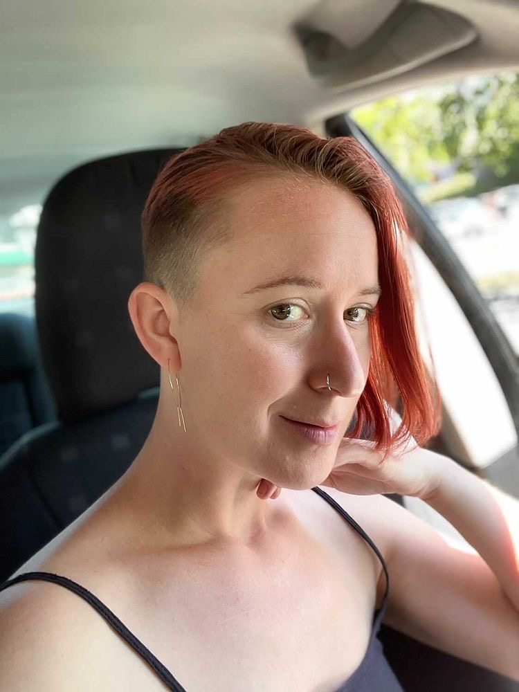 Rachel, a queer pale skinned disabled soul with a nose ring and half a shaved head; the other side has chin length red hair. They are wearing a black cami and sitting in a car, posed with their hand next to their face.
