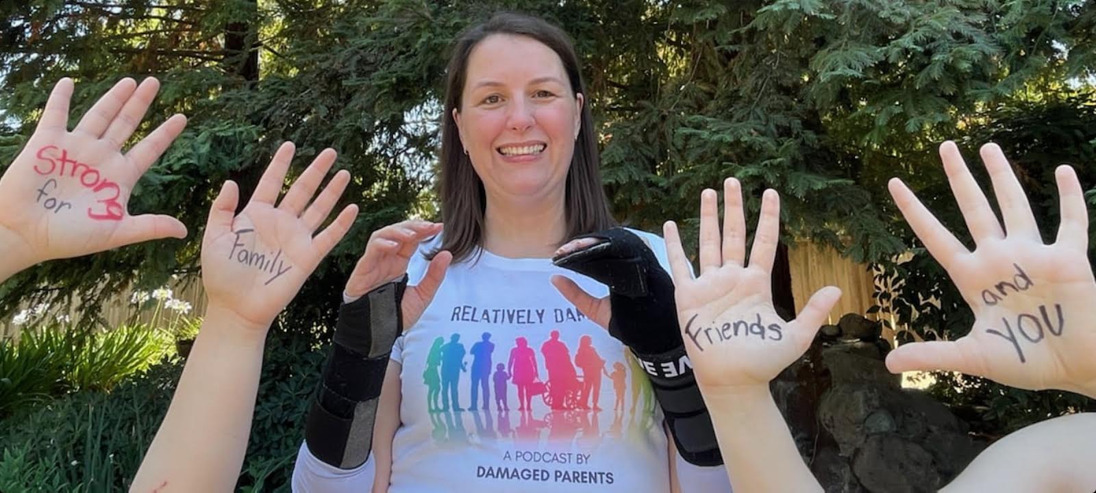 Angela standing in front of redwoods wearing a white t-shirt which says Relatively Damaged with pastel rainbow people of all abilities, shapes and sizes. She is holding up her hands wrapped in black braces with four other hands in picture written on their palms "strong for family friends and you"