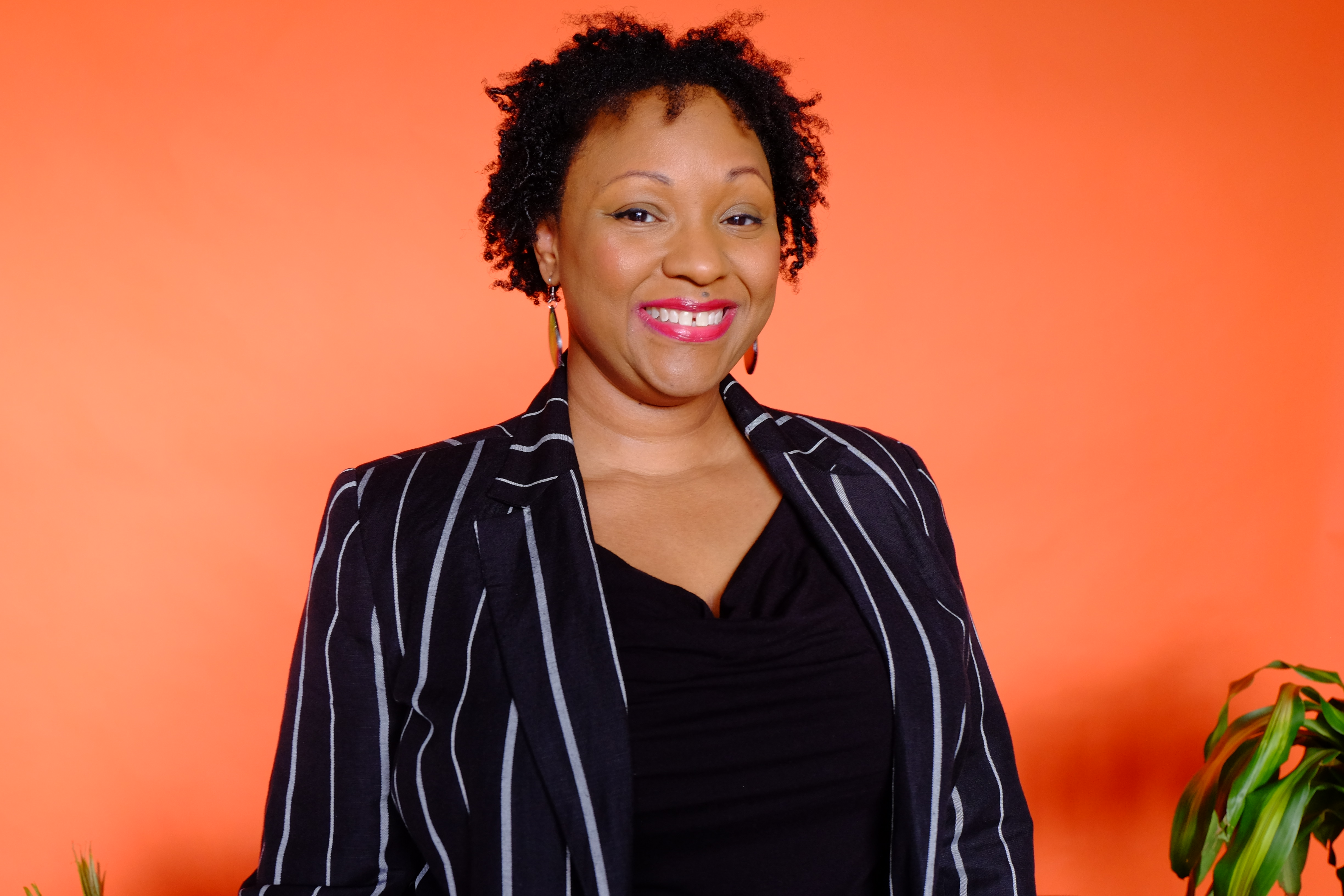 A black woman with curly hair smiling broadly and wearing a black shirt with a black and white striped blazer on an orange background.