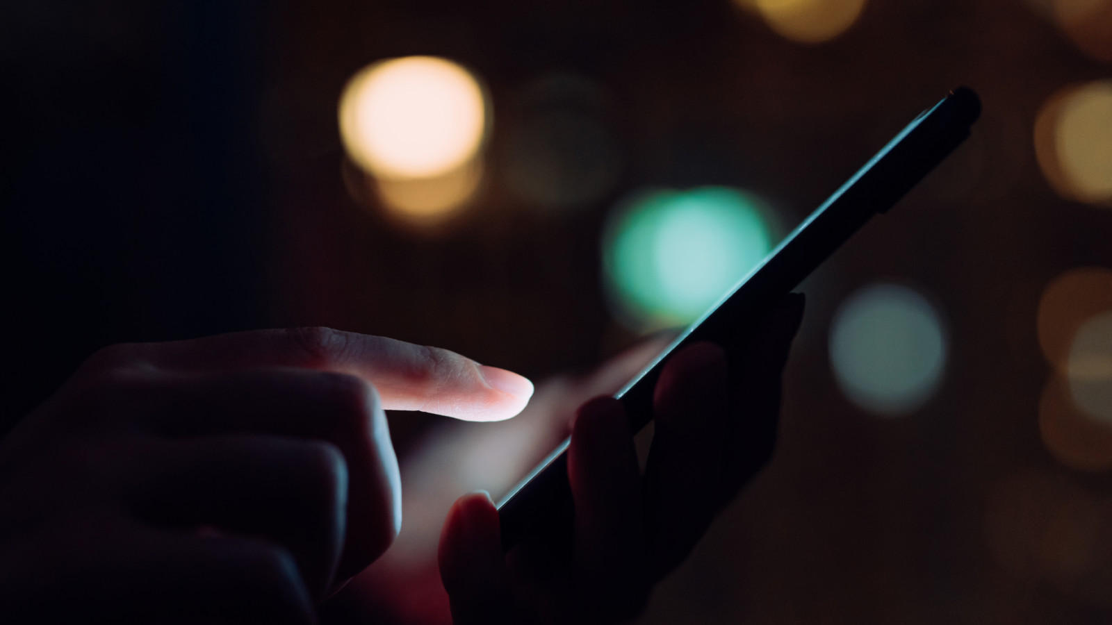Close up of woman's hand using smartphone in the dark, against illuminated city light.