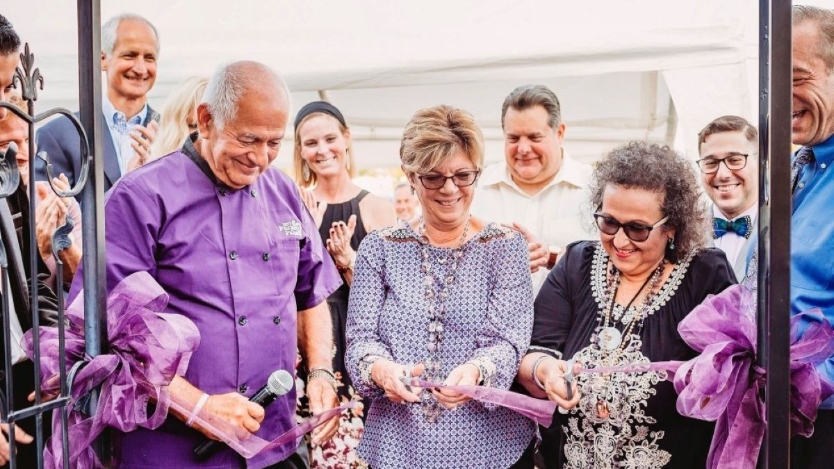 A group of smiling people, with a woman in the center cutting a purple ribbon.