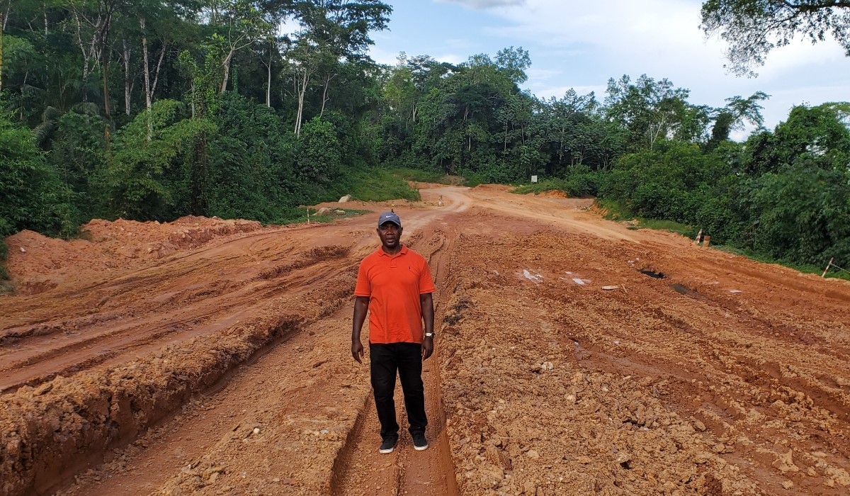 Man in an orange shirt standing on a dirt road with trees in the background.