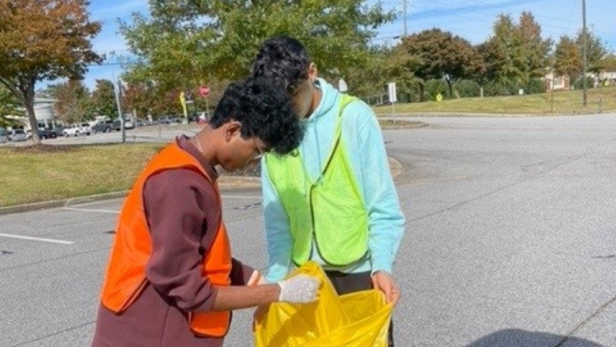 Two people in reflective vests placing trash in a large plastic bag.