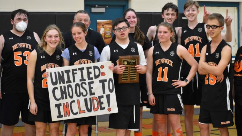 Group picture of a youth basketball team in orange and black uniforms. One player holds a sign reading "Unified: Choose to Include!"
