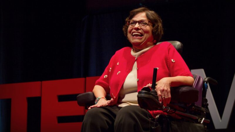 Judy Heumann sitting in her wheelchair, wearing a red sweater. Behind her TED is in large red letters.