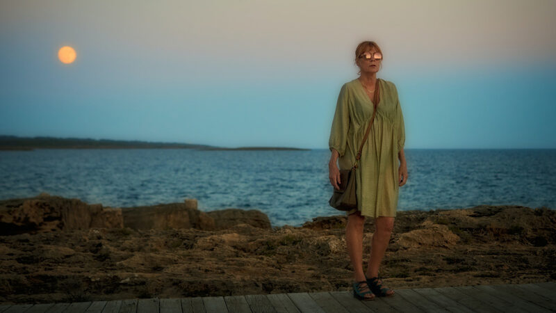A mysterious woman wearing a green dress and sandals faces the viewer, her round sunglasses glinting in the moonlight, as the sky and ocean darken behind her at dusk.
