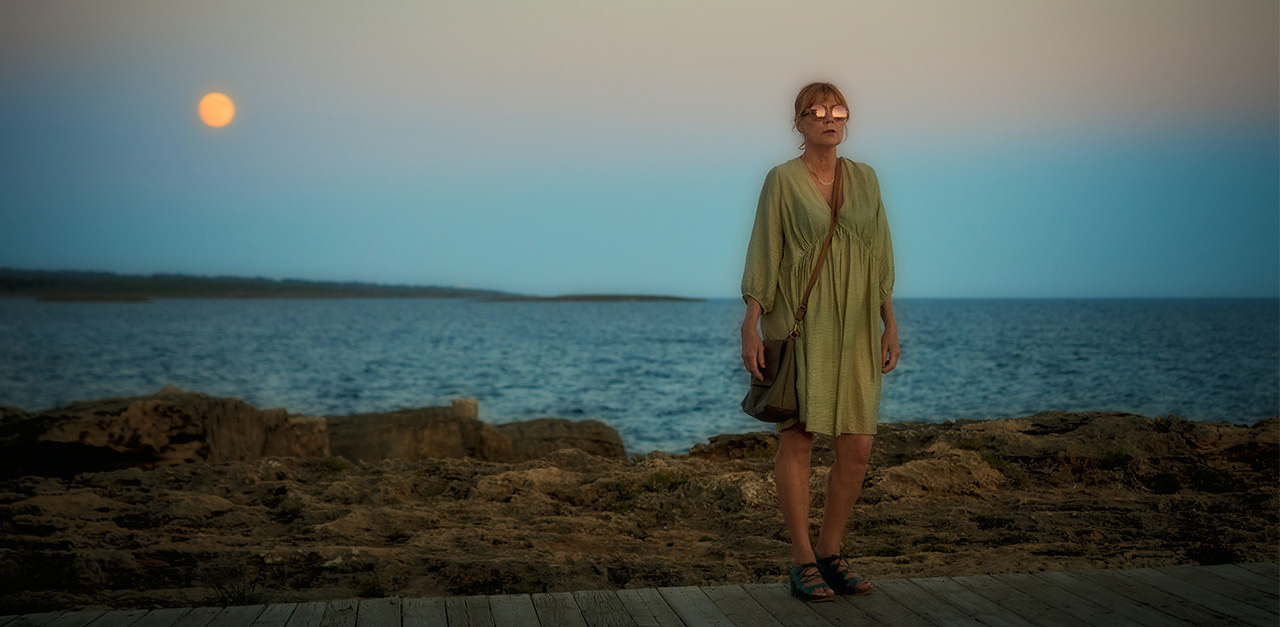 A mysterious woman wearing a green dress and sandals faces the viewer, her round sunglasses glinting in the moonlight, as the sky and ocean darken behind her at dusk.