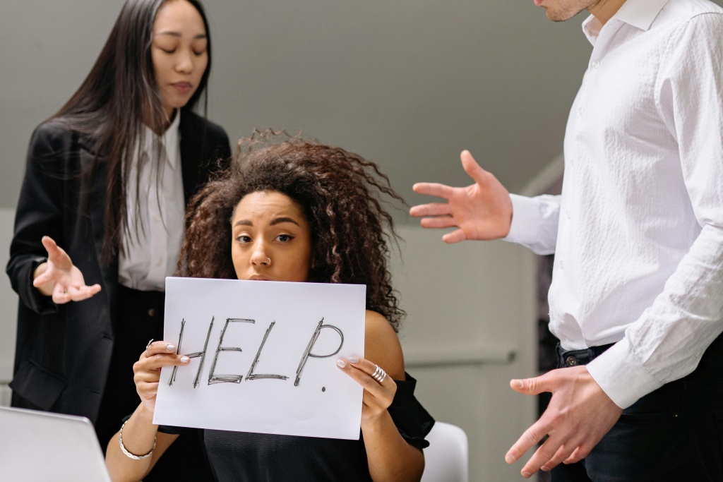 young black woman holding up a help sign while surrounded by coworkers