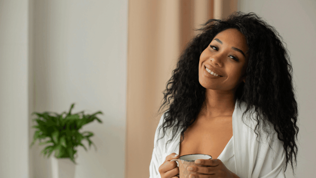 Woman with long curly hair smiling warmly while holding a mug, wearing a white robe, with a green plant blurred in the background.