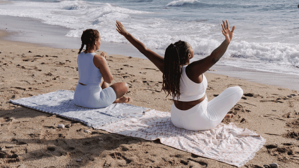 Two women in activewear seated on yoga mats at the beach, facing the ocean, with one raising her arms toward the sky during a mindfulness practice.