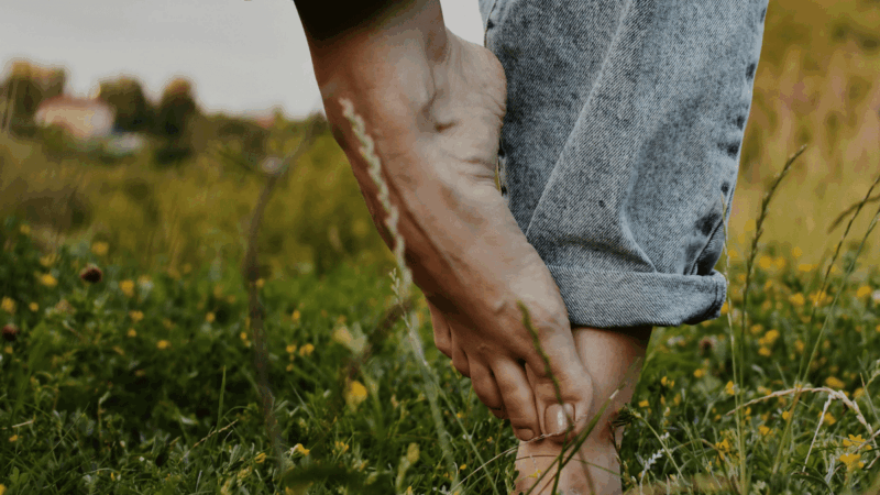 A person stands barefoot in a field of green grass and yellow wildflowers, gently holding their ankle with rolled-up denim pants