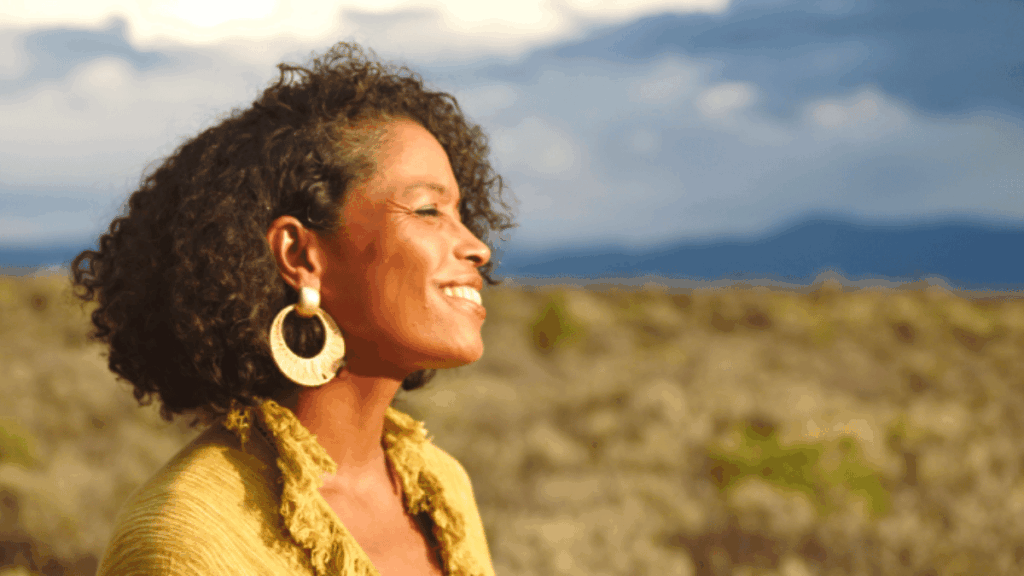 Zee Clarke smiles peacefully in warm sunlight, standing in a desert landscape with mountains and cloudy skies in the distance