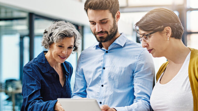 Three colleagues from different generations reviewing information together on a tablet in a modern workplace.