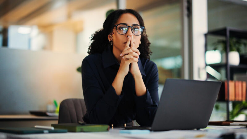 Woman wearing glasses seated at an office desk, looking away from an open laptop with her hands clasped near her face.